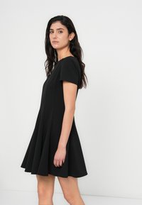 Μικρό μαύρο φόρεμα με κοντά μανίκια, στρογγυλή λαιμόκοψη και φουσκωτή φούστα, που αναδεικνύει ένα λείο ύφασμα και απλή σιλουέτα.