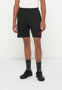Muškarac u crnim Umbro kratkim hlačama, tamnosivim čarapama i crnim Puma sportskim cipelama stoji na bijeloj pozadini.