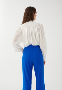 Blusa blanca con mangas abullonadas y pechera fruncida, combinada con pantalones ajustados de un brillante color azul que cuentan con bolsillos traseros y un diseño de pierna recta.
