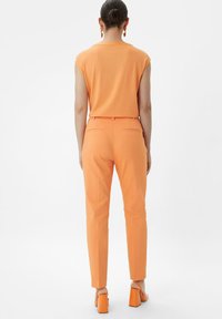 Orange sleeveless top paired with matching tapered trousers; both feature a smooth texture. Notable square-heeled sandals complete the look.