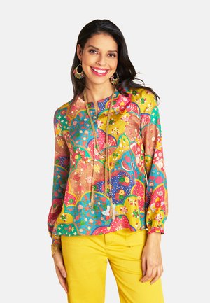 Colorful blouse featuring floral and geometric patterns in pink, yellow, and blue, with long sleeves and a relaxed fit. Paired with yellow pants.