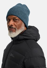 Berretto lavorato a maglia di colore blu scuro con texture a coste. Indossato con una giacca a vento nera con cappuccio e chiusura zip.