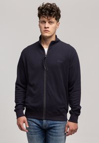 Granatowa kurtka na zamek z teksturowanej tkaniny, z kołnierzem stojącym, zamkiem błyskawicznym i ściągaczami na rękawach, w zestawie z dżinsami.