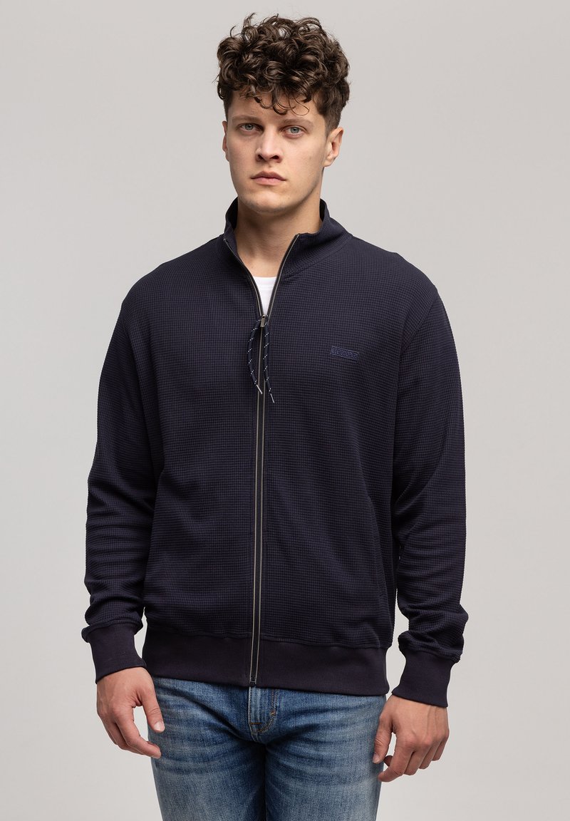 Granatowa kurtka na zamek z teksturowanej tkaniny, z kołnierzem stojącym, zamkiem błyskawicznym i ściągaczami na rękawach, w zestawie z dżinsami.