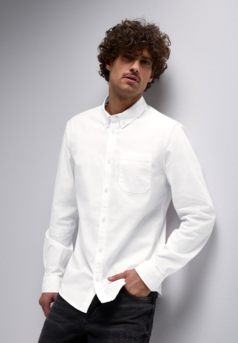 Camisa blanca de botones con un solo bolsillo en el pecho, detalle en el cuello y mangas largas, combinada con jeans oscuros. Diseño limpio y sencillo.