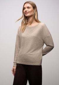 Beiger Pullover mit weitem, rundem Ausschnitt, legerer Passform und gerippten Bündchen, kombiniert mit dunkelbraunen Hosen. Glatte, weiche Textur.