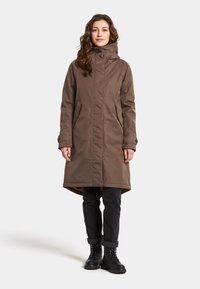 Braune knielange Parka mit hohem Kragen, seitlichen Taschen und Knopfdetails. Getragen über dunklen Jeans und schwarzen Schnürstiefeln.