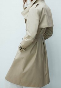 Cappotto trench beige con vita a cintura, bottoni doppi, grande colletto ed epaulettes sulle spalle; di lunghezza media con una texture liscia.
