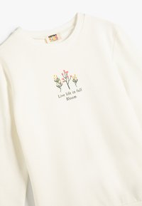 Krämmfärgad sweatshirt i mjuk bomull med rund krage. Har blommönster broderat och texten "Leva livet i full blom."