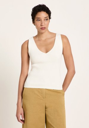 Frau mit kurzen lockigen Haaren, die ein ärmelloses weißes Rippentop und senfgelbe Hose trägt, steht mit einer Hand in der Tasche.