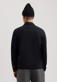 Chaqueta negra con mangas largas, cuello plano y tejido texturizado; cuenta con una yugo trasero y acentos de botones a presión en los puños.