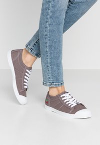 Sneakers in tela grigia con lacci bianchi e punta in gomma. Presentano cuciture bianche e un ricamo di ciliegia rossa sul lato.