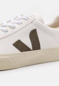 White sneaker with suede olive green V logo, rubber sole, and white laces. Smooth leather upper with texture contrast near logo.