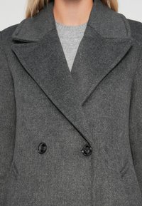 Abrigo gris de doble botonadura con textura de espiga, grandes solapas y botones negros. Superpuesto a un suéter de cuello redondo gris claro.