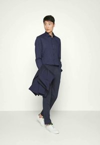 Man met een marineblauw geruit shirt en marineblauwe broek, die een marineblauwe jas vasthoudt, loopt op witte sneakers tegen een effen lichte achtergrond.