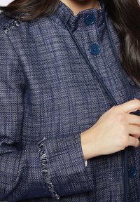 Marinblå jacka i strukturerat tyg med en subtil väv. Har två blå knappar, en hög krage och fransiga manschetter.