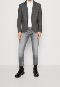 Szary blezer z wciętymi klapami, zapinany na jeden guzik, z przednimi kieszeniami, na białej koszuli, w połączeniu z jasnoszarymi jeansami i czarnymi butami typu combat.