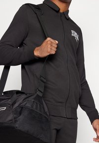 Sudadera negra con cremallera y cuello, con un logo en el pecho, combinada con un bolso de deporte negro que tiene superficies texturizadas y lisas.