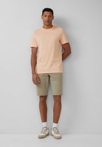 Licht perzik kleurige t-shirt met korte mouwen, beige shorts en crèmekleurige sneakers met bruine accenten. Model staat tegen een grijze achtergrond.
