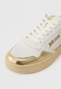 Białe sneakersy z teksturowanym skórzanym wierzchem i złotymi akcentami w wężowy wzór. Posiadają płaskie sznurowadła, okrągły czubek i beżową gumową podeszwę.
