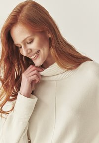 Crème-kleurige trui met een geribbeld col, zachte textuur en een relaxte pasvorm. Het model raakt de stof aan en toont het ontwerp en de kleur.