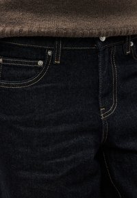 Σκούρο μπλε τζιν με απαλή υφή, με χρυστές ραφές και μπροστινές τσέπες. Η μέση έχει μεταλλικές λεπτομέρειες.