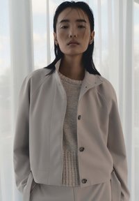 Frau mit glatten dunklen Haaren trägt eine beige Jacke über einem Strickpullover und steht drinnen vor durchscheinenden weißen Vorhängen.