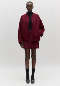 Burgunderfarbene, übergroße Bomberjacke mit Druckknöpfen, kombiniert mit einem passenden, figurbetonten Minirock. Ein schwarzer Rollkragenpullover und klobige schwarze Absätze runden den Look ab.