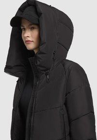 Zwarte pufferjack met een hoge kraag en oversized capuchon, voorzien van een ritssluiting en verstelbare banden, een gladde textuur en minimale details.