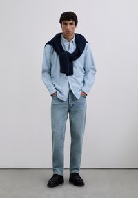 Chemise boutonnée à rayures bleu clair, pull bleu marine noué au cou, jean bleu clair, chaussures noires, debout sur un fond neutre.