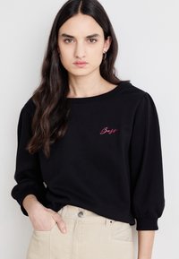 Felpa nera a maniche lunghe con scollo ampio e maniche a palloncino. Presenta un ricamo rosa "Boss" sul davanti. Abbinata a pantaloni beige.