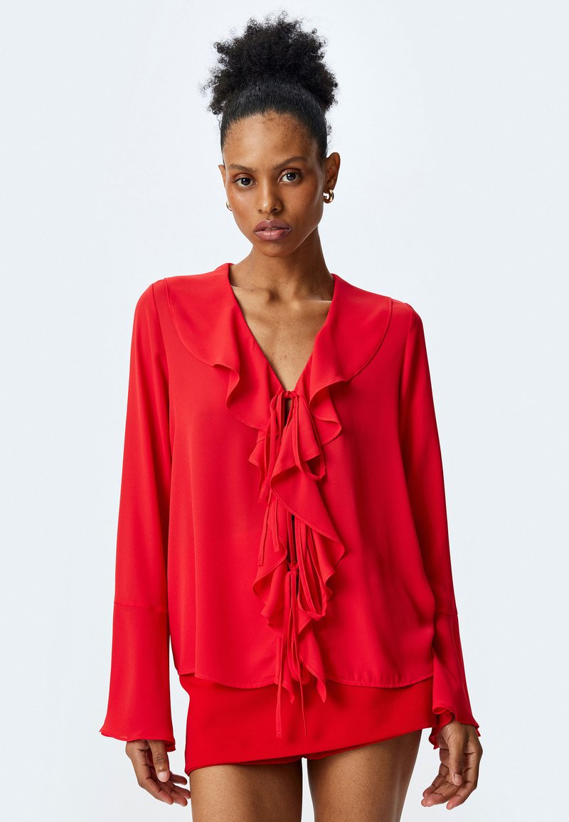 Blusa rossa realizzata in tessuto leggero, con un design anteriore arricciato, scollo a V, maniche lunghe e chiusura a fiocco al collo.