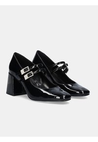 Tacones de charol negro con punta afilada, tacón cuadrado y dos tiras con hebillas de strass cuadradas. Textura suave y acabado brillante.