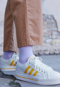 Ljusgrå sneakers med gula ränder, texturerade gummisulor, i kombination med lavendelfärgade strumpor och beige byxor, mot en stenbakgrund.