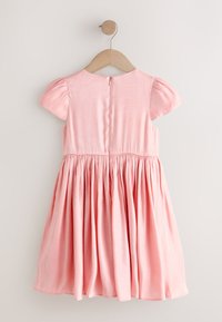 Parte trasera de un vestido rosa claro, de manga corta y cintura fruncida, colgado en una percha de madera sobre un fondo liso.