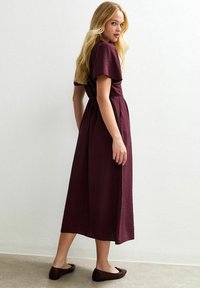 Robe bordeaux à manches courtes, longueur genou, avec une ceinture à nouer à la taille. Portée avec des ballerines marron, texture lisse, design simple.