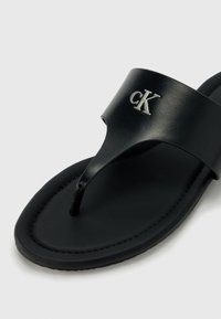 Svart flip-flop sandal med en bred stropp som har et sølvfarget "CK"-logo, glatt tekstur og en konturert fotbed.