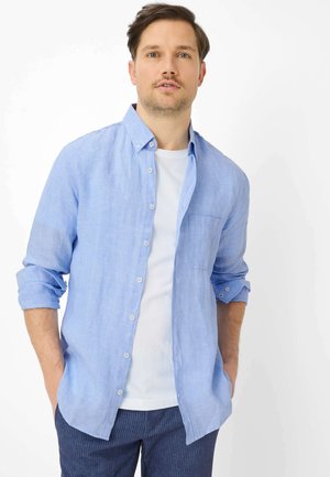 Man draagt een lichtblauw overhemd met knopen over een wit T-shirt en marineblauwe broek met krijtstrepen, handen in de zakken, tegen een witte achtergrond.