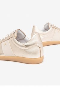 Baskets dorées métalliques avec une texture lisse, des accents blancs et des semelles beige clair. Elles possèdent un laçage classique et un design de bout rond.