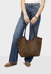Person wearing blue denim jeans and tan flats, holding a large brown leopard print tote bag with dark handles.