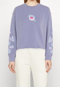 Personne portant un sweat court lavande avec des graphismes en bulles sur les manches et la poitrine, associé à un pantalon blanc.