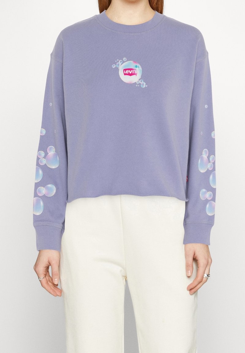 Personne portant un sweat court lavande avec des graphismes en bulles sur les manches et la poitrine, associé à un pantalon blanc.