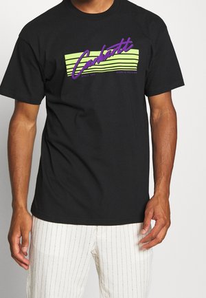 Homme portant un t-shirt noir à manches courtes avec des rayures vert néon et le texte violet "Carhartt", accompagné d'un pantalon blanc à fines rayures.