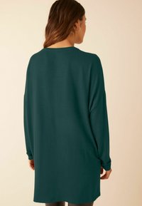 Tunic cu mânecă lungă, supradimensionată, de culoare verde închis, cu guler rotund, textură moale și umeri căzuți, concepută pentru o potrivire lejeră.