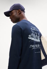 Tumesinine pikkade varrukatega särk, millel on paadielemendid koos aerudega ja figuuridega. Puuvillane materjal, ümar kaelus ja lõdva lõikega. Lihtne seljaosa disain.