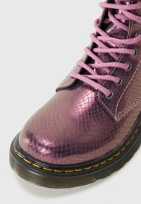 Botín tobillo metálico morado con un patrón de piel de serpiente texturizado, con cordones rosas, ojetes negros y una suela negra con costuras amarillas.