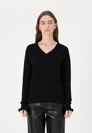 Black v-neck sweater in soft knit fabric, featuring ribbed cuffs with a ruffled edge and a small logo emblem on the chest.