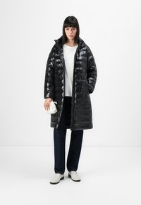 Parka nera imbottita con finitura lucida, cappuccio e design trapuntato. Indossata sopra una camicia bianca e jeans scuri, con una piccola borsa bianca.