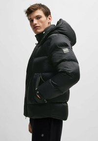 Svart vadderad jacka med en strukturerad yta, som har huva, dragkedjefickor och en märkespatch på vänster ärm.