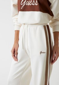 Witte katoenen sweatpants met bruine zijstrepen. Heeft een geborduurd 'Guess' logo en een elastische tailleband met trekkoorden. Zachte textuur.